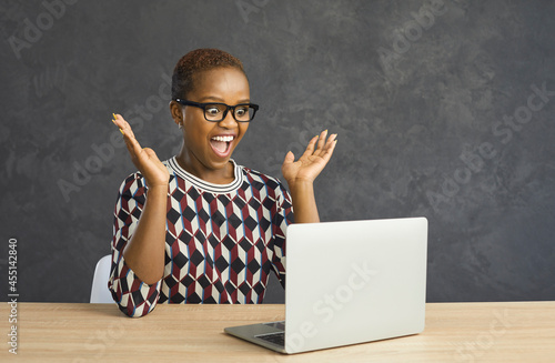 Overjoyed shocked young African American woman in glasses look at laptop screen stunned by win lottery online. Amazed gen z ethnic biracial girl surprised by unexpected good news on computer on web.