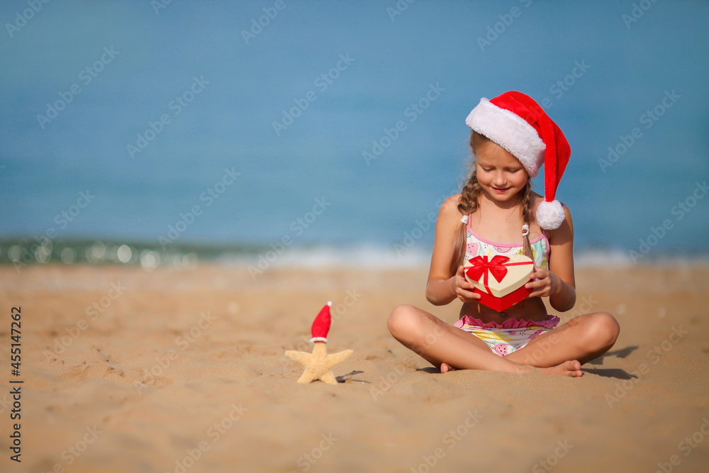 Obraz premium The child sits on the sand wearing a Santa hat. Little girl smiles and looks at a box with a gift. Merry Christmas. Winter vacation. Sea in the background. Copy space.