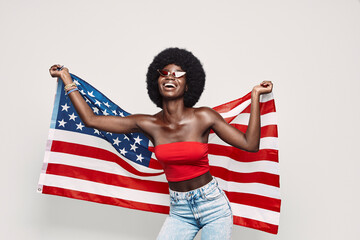 Happy young African woman carrying American flag and smiling while standing against gray background