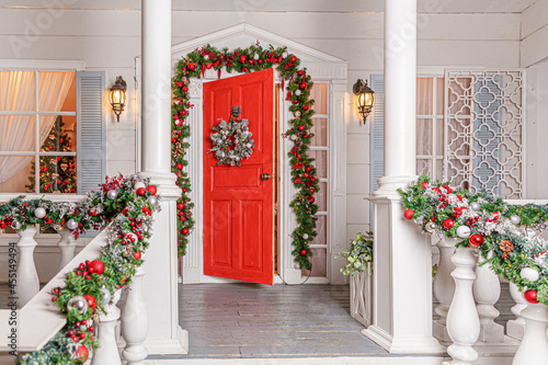 Christmas porch decoration idea. House entrance with red door decorated for holidays. Red and green wreath garland of fir tree branches and lights on railing. Christmas eve at home