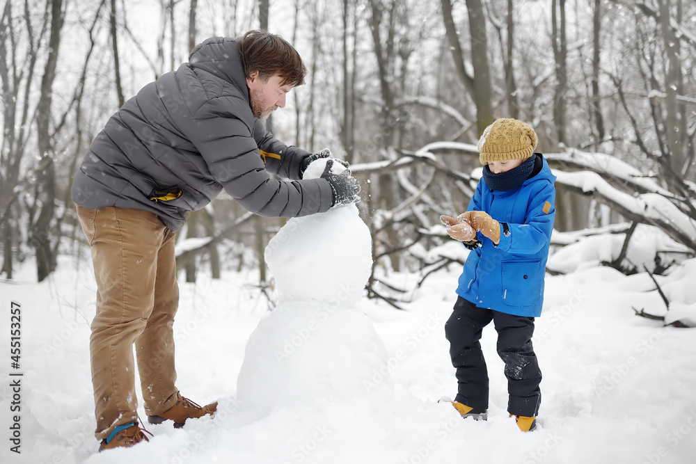 Little boy with his father building snowman in snowy park. Active outdoors leisure with children in winter. Kid during stroll in a snowy winter park