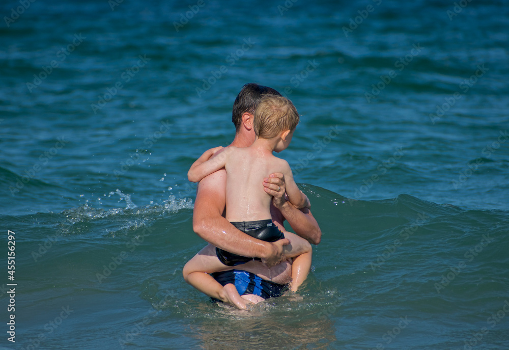 A young father with his child in his arms in the waves of the sea. Parents' care for children - concept. Rear view.