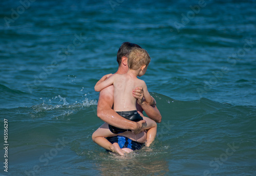 A young father with his child in his arms in the waves of the sea. Parents' care for children - concept. Rear view.