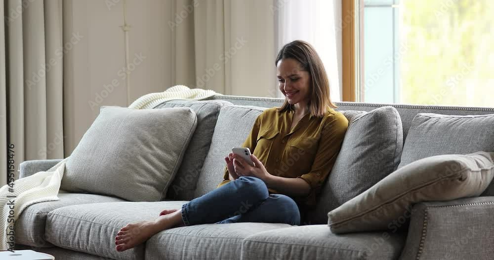 Attractive young woman sit on sofa in living room with smartphone, social media user chatting on-line, writing messages, share news with friend, communicates remotely. Modern tech, lifestyle concept Attractive young woman sit on sofa in living room with smartphone, social media user chatting on-line, writing messages, share news with friend, communicates remotely. Modern tech, lifestyle concept