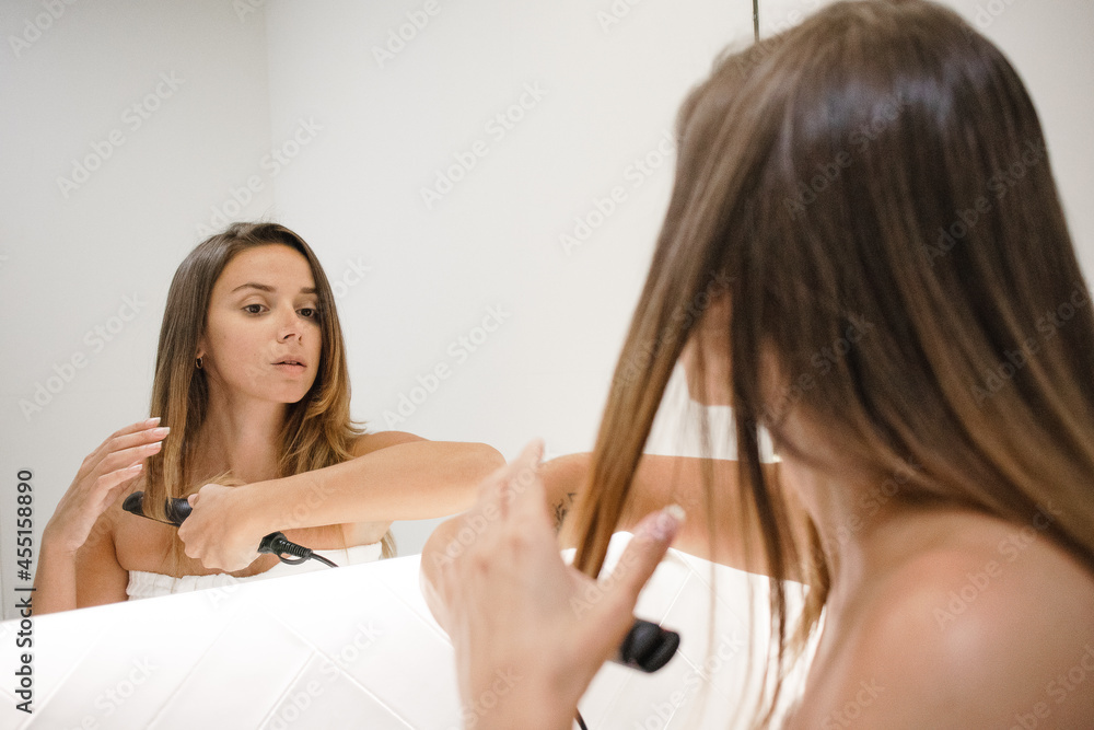 young beautiful millennial dark blond brunette white woman at home no makeup long hair after shower looking at herself in the bathroom mirror using hair straightener self care routine self love