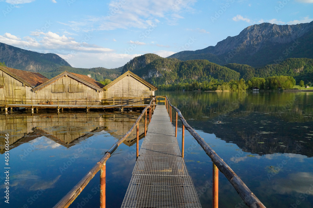 a pier leading to the wooden boat houses on tranquil lake Kochel (Kochelsee) with the scenic Bavarian Alps in the background reflected in the water (Bavaria, Germany)	