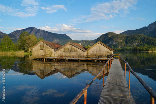 pier leading to the wooden boat houses on tranquil lake Kochelsee with the scenic Bavarian Alps and blue sky in the background reflected in the clear waters of the alpine lake (Bavaria, Germany) 