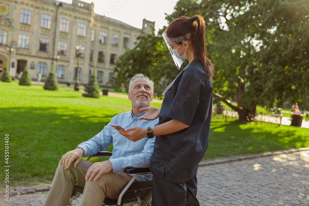 Cheerful mature man in wheelchair smiling at young nurse wearing face shield and mask, on a walk together outdoors Cheerful mature man in wheelchair smiling at young nurse wearing face shield and mask, on a walk together outdoors