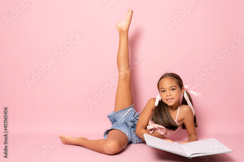 Fototapeta Naklejka Na Ścianę i Meble -  Beautiful little girl lying on the floor with one leg up studying isolated over pink background.