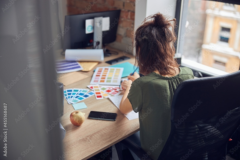 Back view of interior designer with messy hairdo making notes in her notebook, selecting color samples, working on creative project in her office Back view of interior designer with messy hairdo making notes in her notebook, selecting color samples, working on creative project in her office