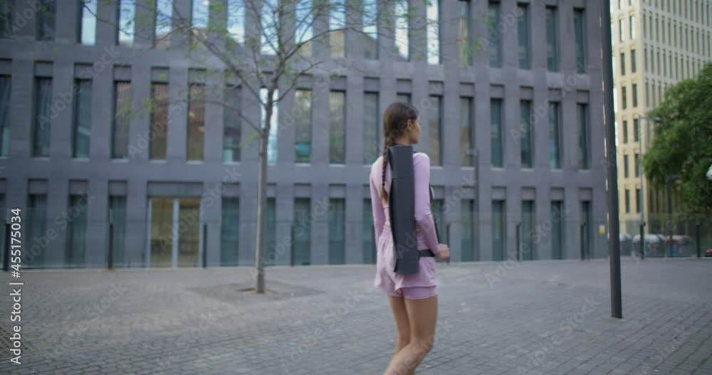 Slim active young woman walks around city has pilates exercisess with fitness mat passes by modern building walks to sports club alone covers long distance per day goes to location for workout Slim active young woman walks around city has pilates exercisess with fitness mat passes by modern building walks to sports club alone covers long distance per day goes to location for workout