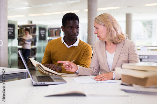 Friendly female tutor helping to diligent african-american man preparing for exam in library. High quality photo © JackF