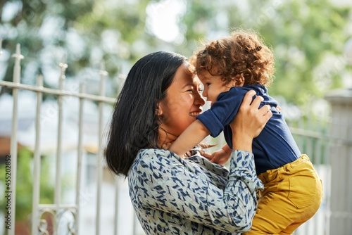 Joyful mother and little son spending time together, parent lifting her baby when playing outdoors