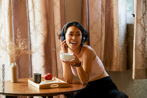 Happy excited young woman enjoying good music or podcast in headphone and eating tasty breakfast at home