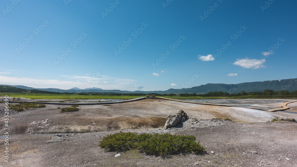 Fototapeta premium The caldera of an extinct volcano has rocky soil. Plank paths are laid for tourists. In the distance there is a green meadow. A mountain range against the blue sky. Kamchatka. Uzon