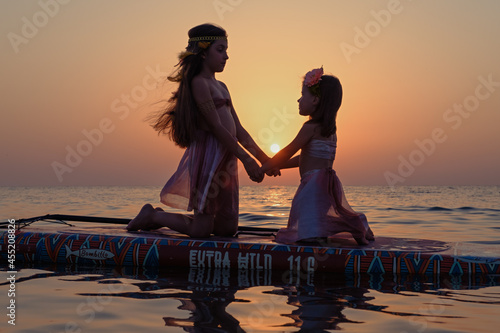 Two girls in the image of Moana on a boat in the sea in the rays of the sunset.