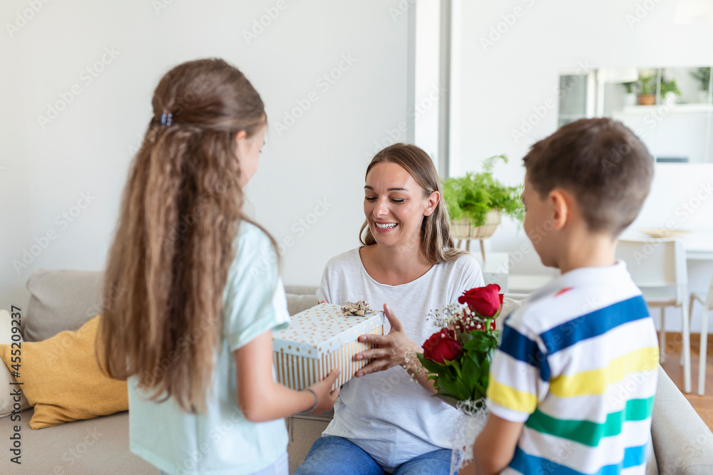 Happy children giving gift an flowers to mother. Happy Mothers Day! Children boy and girl congratulate smiling mother, give her flowers bouquet of roses and a gift box during holiday celebration Happy children giving gift an flowers to mother. Happy Mothers Day! Children boy and girl congratulate smiling mother, give her flowers bouquet of roses and a gift box during holiday celebration
