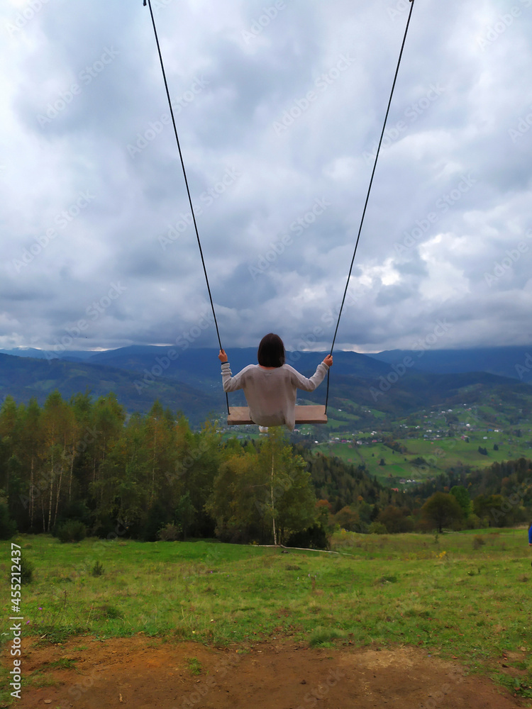 Back of girl with dark hair in a gray sweater sitting on a swing with the mountain view. Calm and quiet wanderlust concept moment when person feels happiness and life. Alone travel in Ukraine.