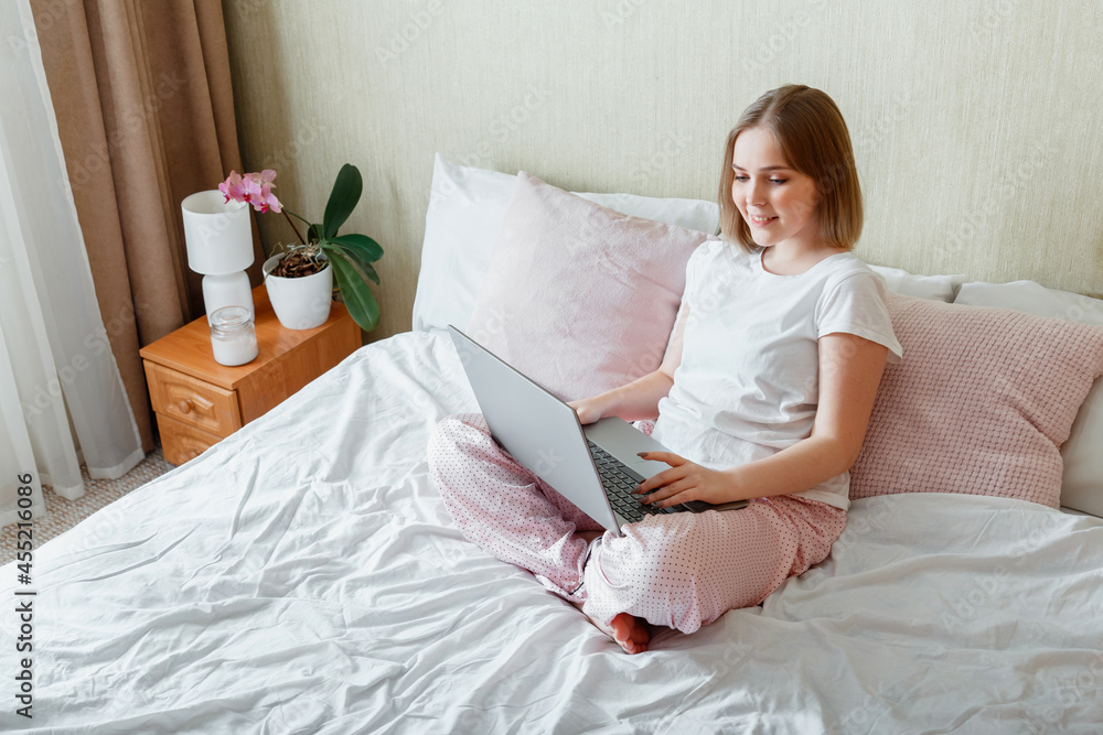 Young woman smile and work using laptop while sitting in bed at morning home. Happy girl in pajamas studying online shopping or planning her day use computer laptop in bedroom interior. Young woman smile and work using laptop while sitting in bed at morning home. Happy girl in pajamas studying online shopping or planning her day use computer laptop in bedroom interior.