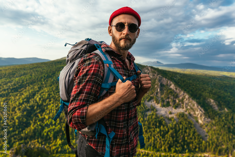 A bearded traveler with a backpack on the top of a mountain. Portrait of a traveler in a red cap and sunglasses. A tourist with a backpack stands against the background of a mountain. Copy space A bearded traveler with a backpack on the top of a mountain. Portrait of a traveler in a red cap and sunglasses. A tourist with a backpack stands against the background of a mountain. Copy space