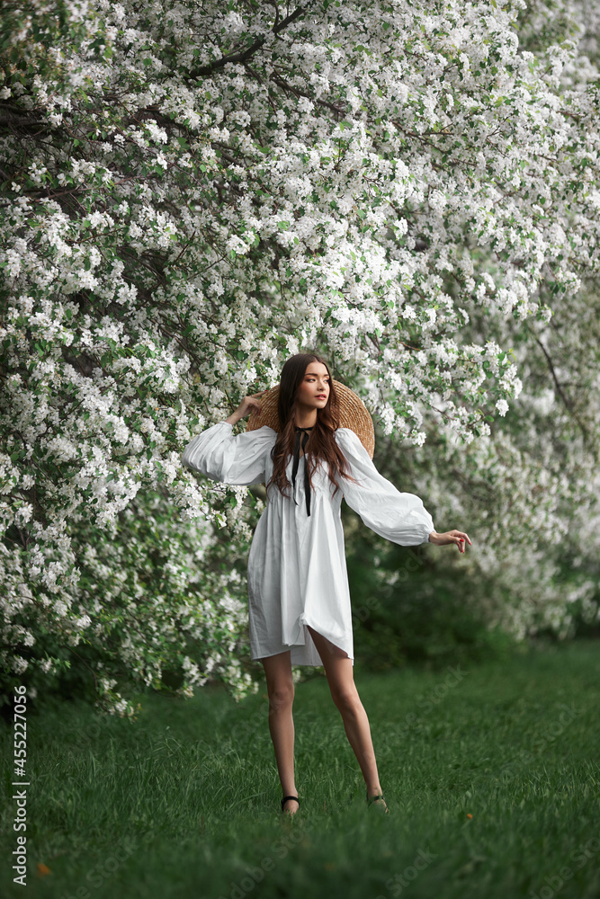 Young woman in white dress with a straw hat walks through blooming spring garden park. Spring has come, romantic mood Young woman in white dress with a straw hat walks through blooming spring garden park. Spring has come, romantic mood