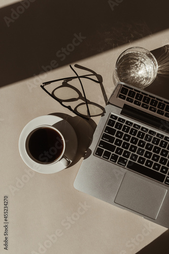 Home office workspace with laptop computer, coffee cup, glasses, crystal glass in sunlight shadows. Flat lay, top view aesthetic luxury bohemian minimal lady boss business lifestyle concept