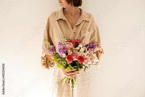 Schilderij op canvas Young beautiful woman in beige muslin shirt hold bouquet of colourful roses flowers against white wall