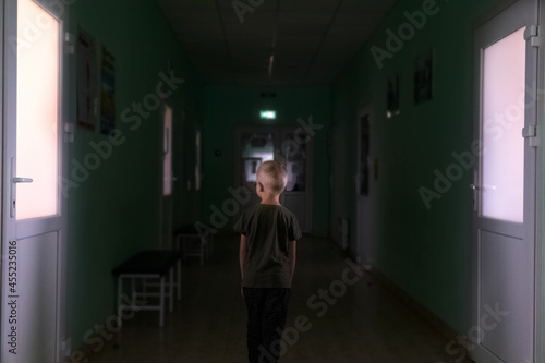 The child stands in the middle of the dark corridor of the hospital. Clinic fear and panic attacks. The boy is afraid of doctors. Conceptual photo of infant panic anxiety. Poorly lit place