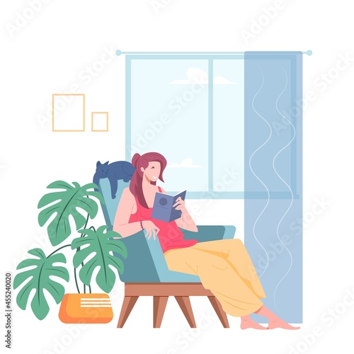 Young woman reading book while sitting in chair in room near window. Pastime scene by young woman in home interior.