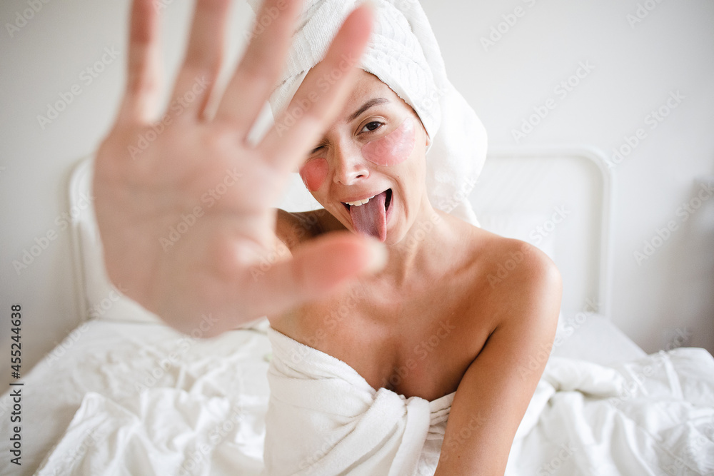 young white carefree woman no makeup sitting on the bed self care with pink under eye patches starring funnily at camera playfully showing tongue using her hand to hide from camera self love happy fun young white carefree woman no makeup sitting on the bed self care with pink under eye patches starring funnily at camera playfully showing tongue using her hand to hide from camera self love happy fun