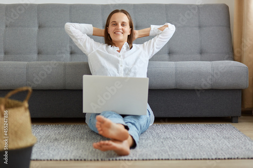 Young adult happy confident woman wearing white shirt and jeans sitting with notebook on legs  raised arms behind head  looking at camera  freelancer being satisfied with finished work.