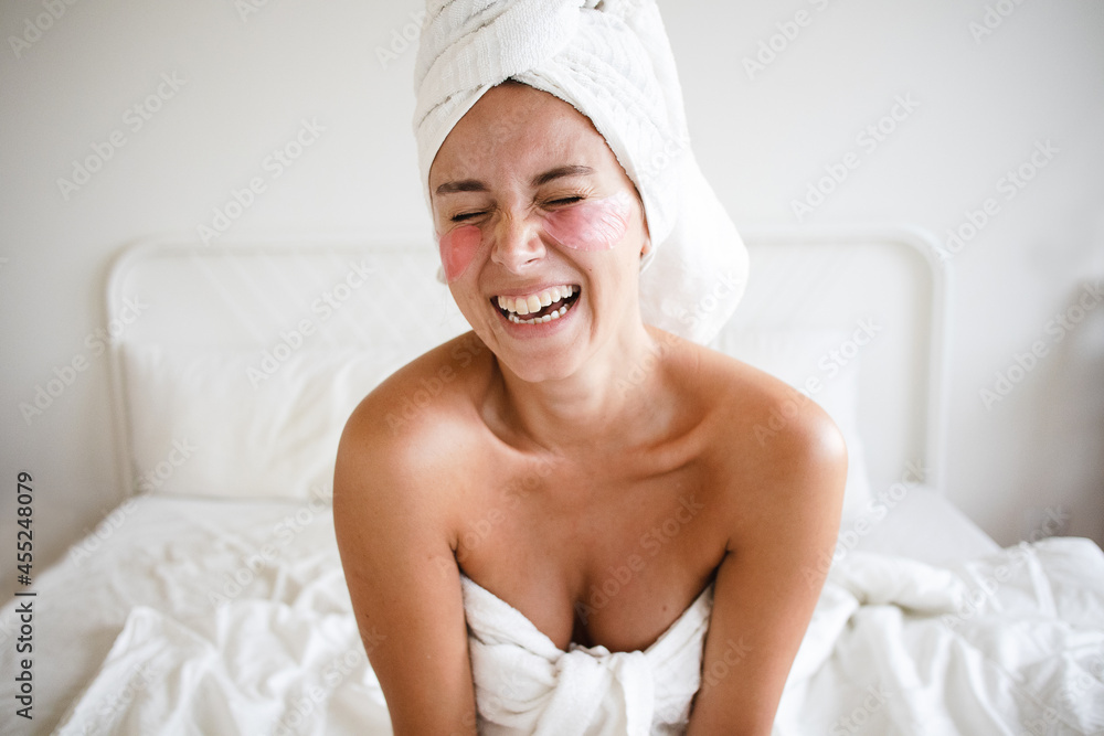 young woman self-care routine no makeup sitting on the bed with pink under-eye patches laughing having fun using hand to hide from camera towel on her head self love happy fun mental health joy white