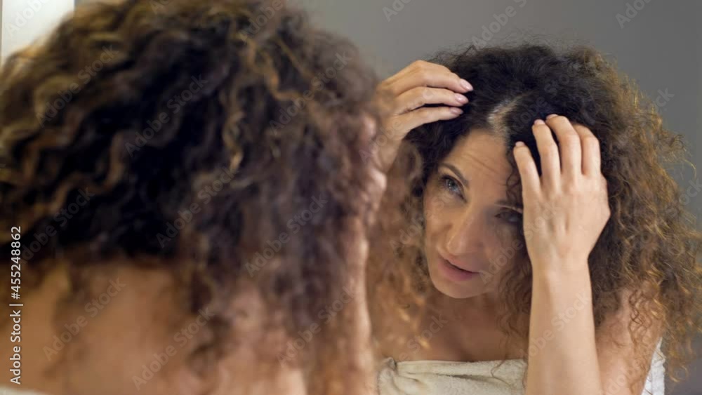 Beautiful middle-aged woman examines her gorgeous hair in the mirror. She cares about gray hair. Beautiful middle-aged woman examines her gorgeous hair in the mirror. She cares about gray hair.
