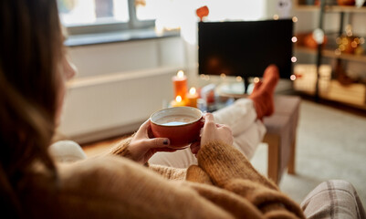 halloween, holidays and leisure concept - young woman watching tv and drinking hot chocolate with her feet on table at cozy home