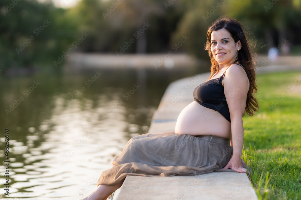 Smiling pregnant woman sitting on the edge of a river in a park during sunset Smiling pregnant woman sitting on the edge of a river in a park during sunset