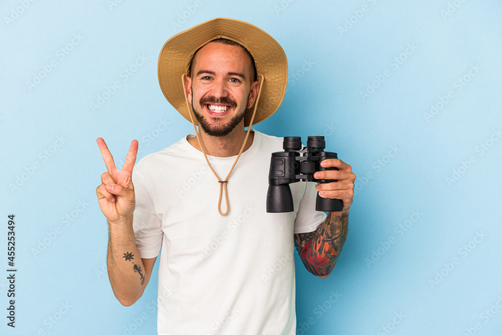 Young caucasian man with tattoos holding binoculars isolated on blue background showing number two with fingers. Young caucasian man with tattoos holding binoculars isolated on blue background showing number two with fingers.