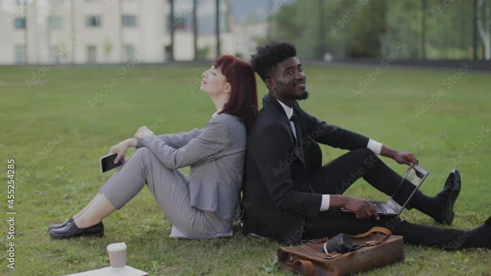 Relaxation after hard work, well done. Close up of multiethnic two business office colleagues, sitting back to back on the grass and relaxing during break outdoors Relaxation after hard work, well done. Close up of multiethnic two business office colleagues, sitting back to back on the grass and relaxing during break outdoors