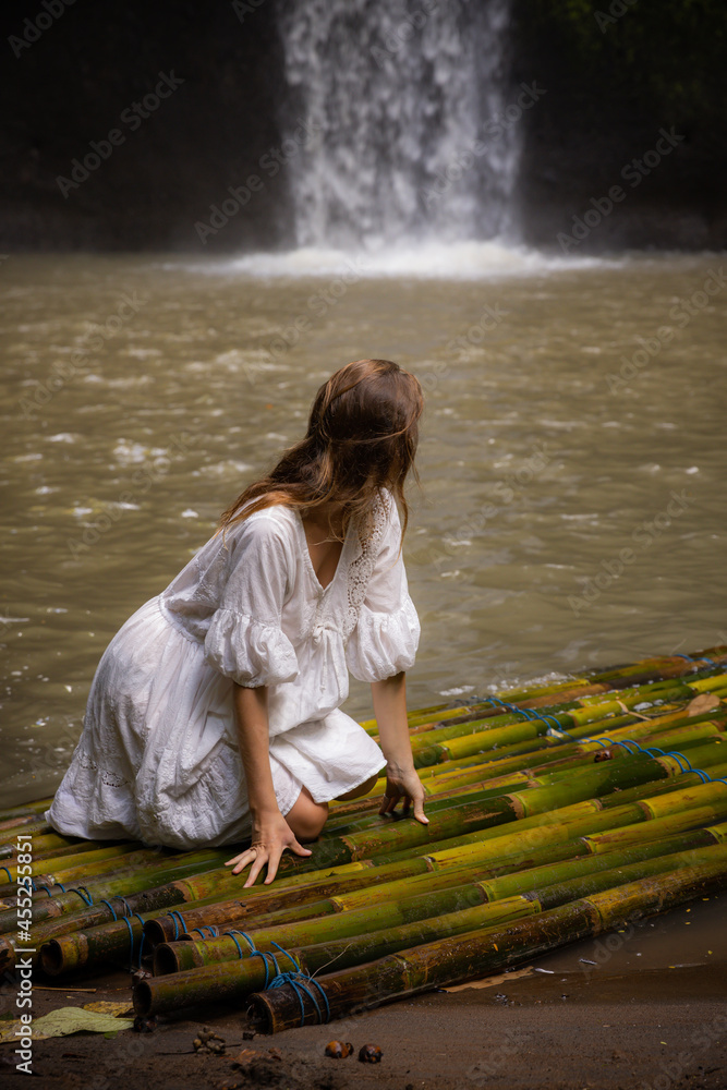 Young woman sitting on bamboo raft. Beautiful Caucasian woman wearing white dress. Travel lifestyle. Nature and environment concept. Tibumana waterfall, Bali