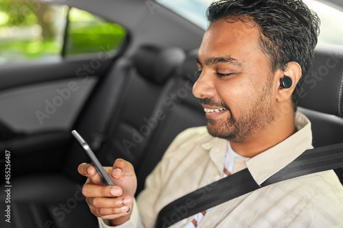 transportation, technology and people concept - happy smiling indian male passenger with wireless earphones using smartphone on back seat of taxi car