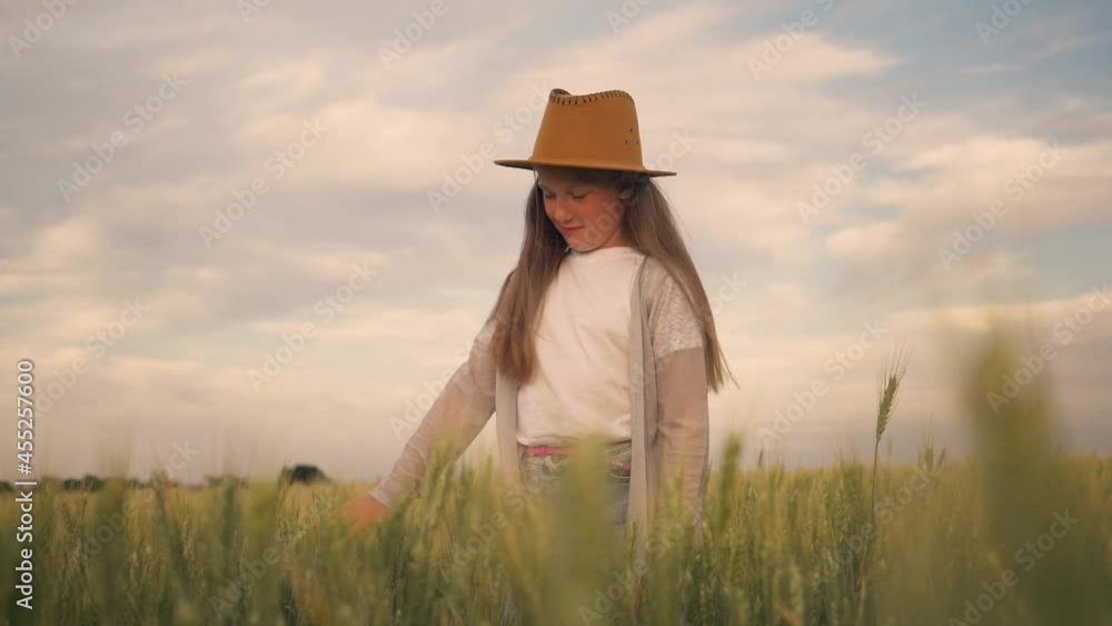 A happy child in a hat in a green wheat field holding a spikelet. A girl in a grain field looks at the harvest of organic products. The girl farmer enjoys the richness and abundance of the crop. A happy child in a hat in a green wheat field holding a spikelet. A girl in a grain field looks at the harvest of organic products. The girl farmer enjoys the richness and abundance of the crop.