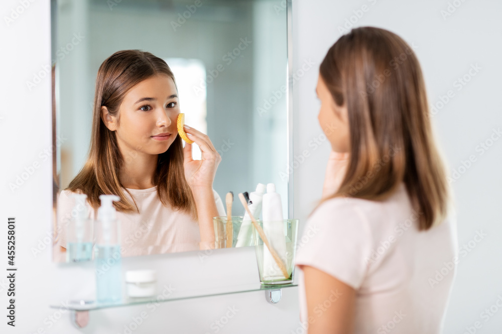 beauty, hygiene and people concept - teenage girl with cleansing sponge cleaning facial skin and looking in mirror at bathroom beauty, hygiene and people concept - teenage girl with cleansing sponge cleaning facial skin and looking in mirror at bathroom