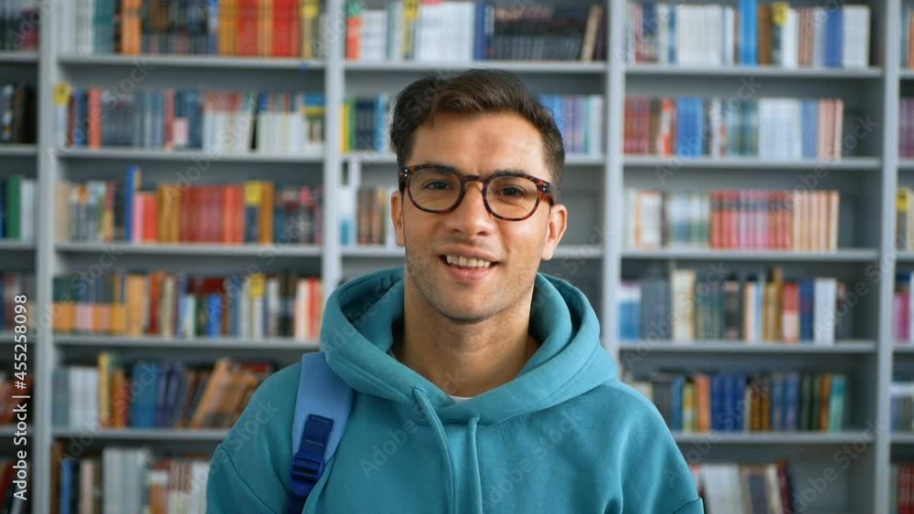 Young student millennial with glasses looking at the camera and smiling while standing in a public library against the background of bookshelves. Close-up portrait of a young attractive guy student Young student millennial with glasses looking at the camera and smiling while standing in a public library against the background of bookshelves. Close-up portrait of a young attractive guy student