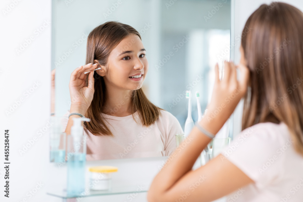 beauty, hygiene and people concept - teenage girl looking in mirror and cleaning ear with cotton swab at bathroom beauty, hygiene and people concept - teenage girl looking in mirror and cleaning ear with cotton swab at bathroom