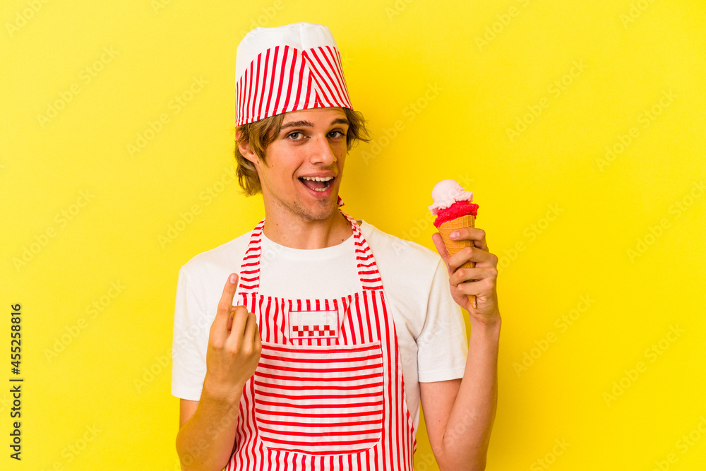 Young ice cream maker man with makeup holding ice cream isolated on yellow background pointing with finger at you as if inviting come closer. Young ice cream maker man with makeup holding ice cream isolated on yellow background pointing with finger at you as if inviting come closer.