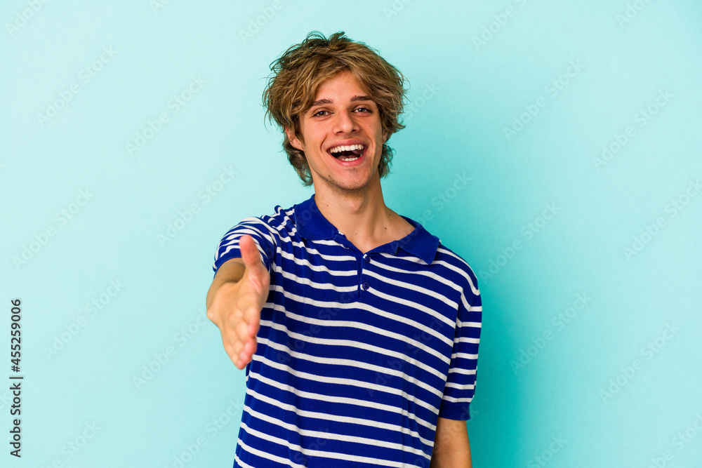 Young caucasian man with make up isolated on blue background stretching hand at camera in greeting gesture. Young caucasian man with make up isolated on blue background stretching hand at camera in greeting gesture.