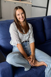 Portrait of serene and charming beautiful dark-haired woman. Pretty caucasian lady in casual wear looks at the camera and smiles, sitting on the comfortable sofa in modern apartment