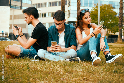 Three friends sitting in the grass in the park and looking at their phones.