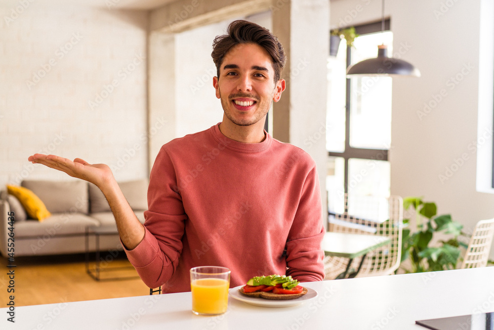 Young mixed race man having breakfast in his kitchen showing a copy space on a palm and holding another hand on waist. Young mixed race man having breakfast in his kitchen showing a copy space on a palm and holding another hand on waist.