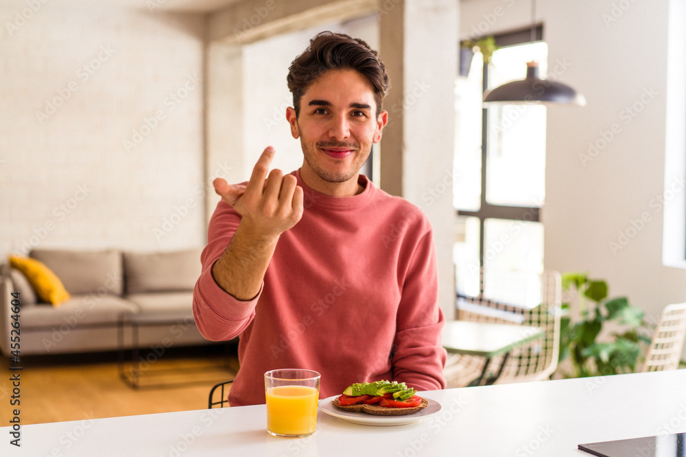 Young mixed race man having breakfast in his kitchen pointing with finger at you as if inviting come closer. Young mixed race man having breakfast in his kitchen pointing with finger at you as if inviting come closer.