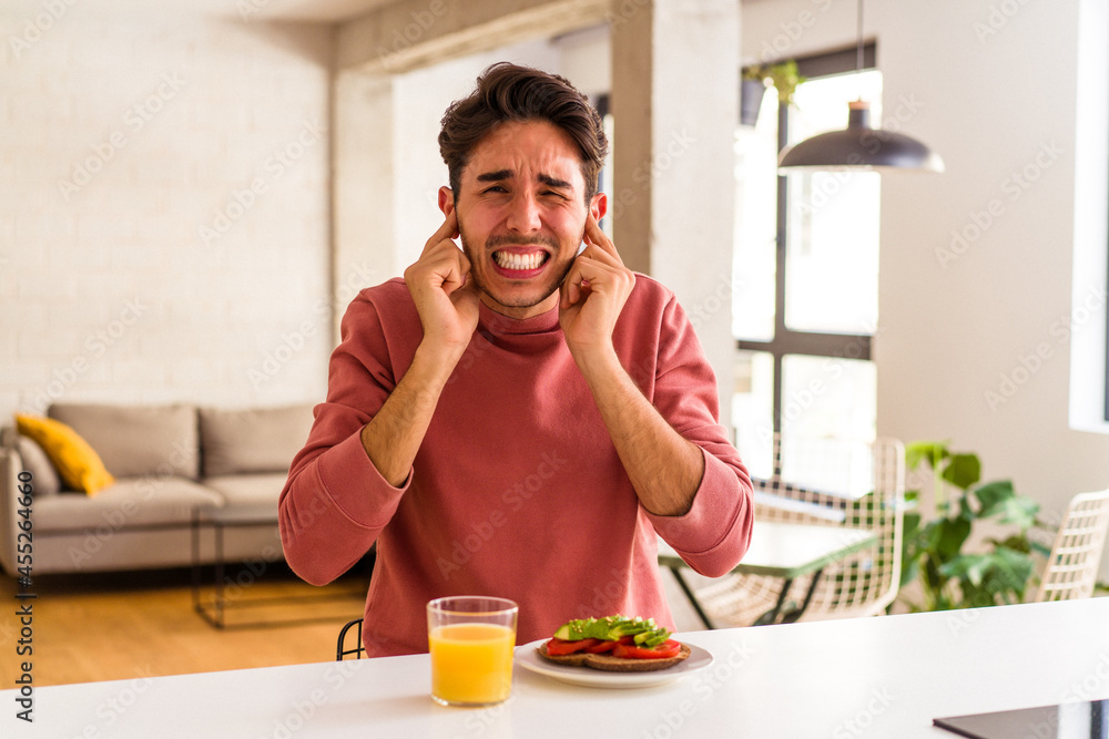 Young mixed race man having breakfast in his kitchen covering ears with hands. Young mixed race man having breakfast in his kitchen covering ears with hands.